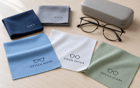 Gamuzas de microfibra extendidas y dobladas con logotipos de ópticas junto a unas gafas sobre una mesa clara.
